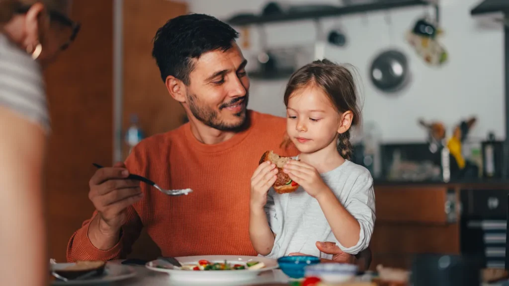 Pai e filha almoçando juntos em casa, com a menina segurando um pedaço de comida e o pai sorrindo enquanto usa o talher à mesa. Ambiente aconchegante e familiar.