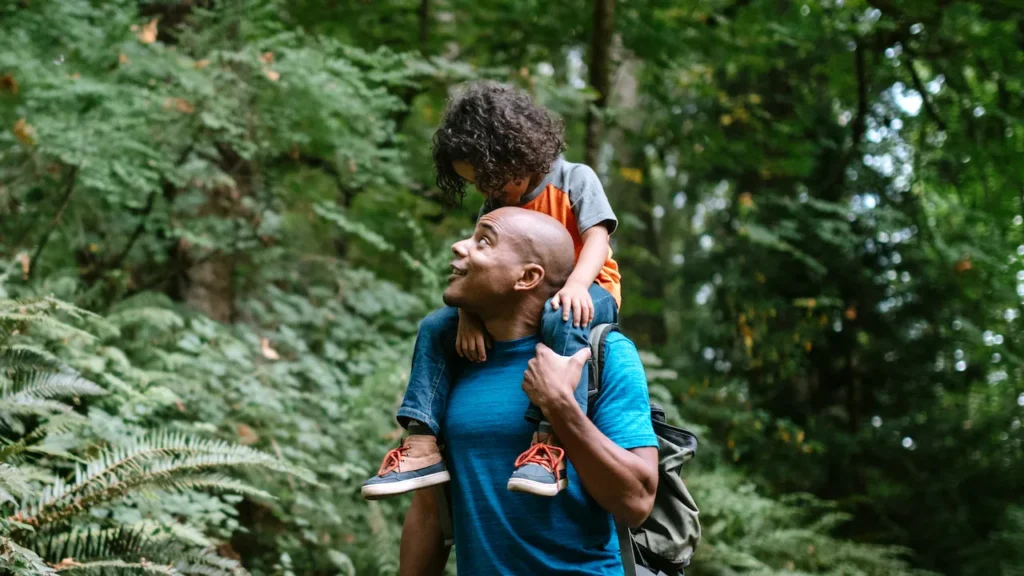 Pai carregando uma criança pequena no colo em meio à natureza, em um ambiente verde com árvores e plantas ao fundo, demonstrando carinho e cuidado ao ar livre.